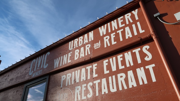 A building facade with bold white lettering on a reddish-brown wall advertising an urban winery, wine bar, restaurant, and space for private events. The text is prominently displayed against the backdrop of a clear blue sky.