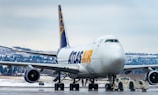 A large commercial cargo airplane is stationed on an airport runway, showing the front and side view. The aircraft is branded with 'ATLAS AIR' in bold yellow and blue letters. Snow-covered terrain and evergreen trees can be seen in the background, indicating a cold, wintry setting. A small airport vehicle is positioned under the plane's nose, hinting at aircraft maintenance or towing activity.