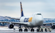 A large commercial cargo airplane is stationed on an airport runway, showing the front and side view. The aircraft is branded with 'ATLAS AIR' in bold yellow and blue letters. Snow-covered terrain and evergreen trees can be seen in the background, indicating a cold, wintry setting. A small airport vehicle is positioned under the plane's nose, hinting at aircraft maintenance or towing activity.