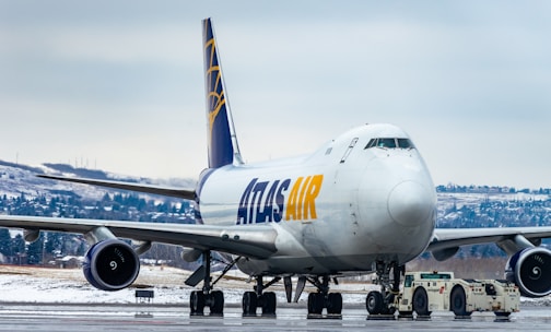 A large commercial cargo airplane is stationed on an airport runway, showing the front and side view. The aircraft is branded with 'ATLAS AIR' in bold yellow and blue letters. Snow-covered terrain and evergreen trees can be seen in the background, indicating a cold, wintry setting. A small airport vehicle is positioned under the plane's nose, hinting at aircraft maintenance or towing activity.