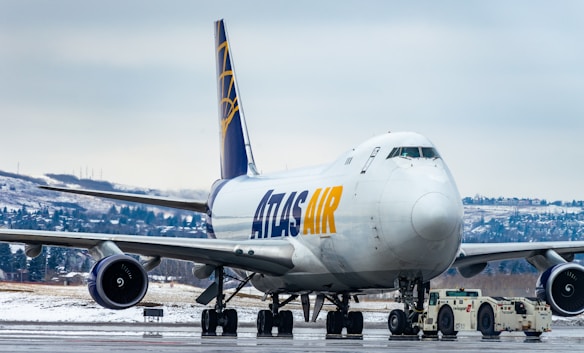 A large commercial cargo airplane is stationed on an airport runway, showing the front and side view. The aircraft is branded with 'ATLAS AIR' in bold yellow and blue letters. Snow-covered terrain and evergreen trees can be seen in the background, indicating a cold, wintry setting. A small airport vehicle is positioned under the plane's nose, hinting at aircraft maintenance or towing activity.