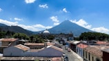 A picturesque town with colonial architecture and a prominent central dome is set against the backdrop of a large, cone-shaped volcano under a bright blue sky with scattered clouds.