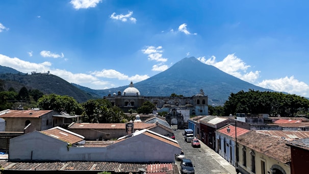 A picturesque town with colonial architecture and a prominent central dome is set against the backdrop of a large, cone-shaped volcano under a bright blue sky with scattered clouds.