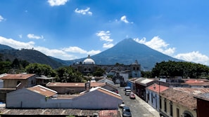 A picturesque town with colonial architecture and a prominent central dome is set against the backdrop of a large, cone-shaped volcano under a bright blue sky with scattered clouds.