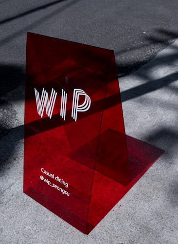 A translucent red acrylic sign with the letters 'WIP' prominently displayed. Below, the text reads 'Casual dining @wip_seongsu'. The sign casts a shadow on the concrete pavement, which is partially lit by sunlight.