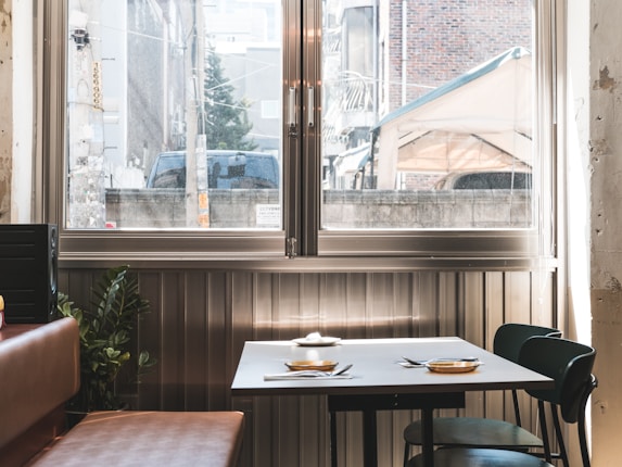A table set for two is positioned next to a window in a cozy, softly lit room. The table has simple tableware, including plates and cutlery. Outside the window, an urban scene is visible with buildings and a parked vehicle.