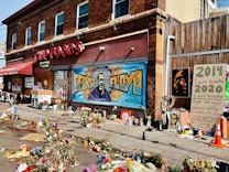 A mural of George Floyd is painted on the wall of a building that says CUP FOODS. The area in front is filled with flowers and memorials. A large poster displays the years 2014 and 2020, with messages about not being able to breathe and a call for change. Various items like traffic cones are scattered around.