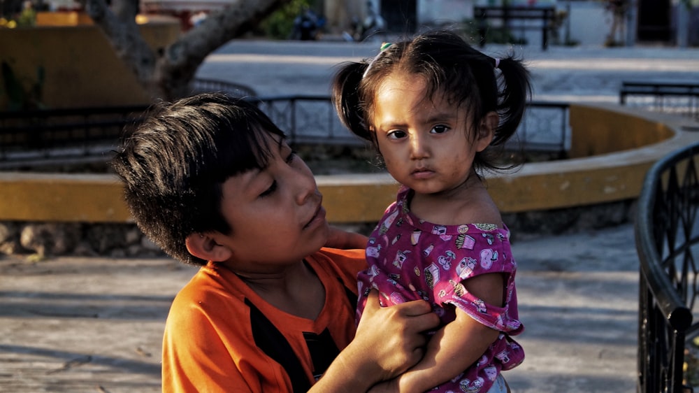 boy in orange crew neck shirt carrying girl in pink and white floral shirt