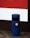 A blue cylindrical outdoor rubbish bin with a yellow label is situated on a paved sidewalk. It's positioned against a building featuring a section of red and white painted panels.