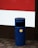 A blue cylindrical outdoor rubbish bin with a yellow label is situated on a paved sidewalk. It's positioned against a building featuring a section of red and white painted panels.