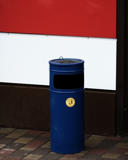 A blue cylindrical outdoor rubbish bin with a yellow label is situated on a paved sidewalk. It's positioned against a building featuring a section of red and white painted panels.