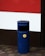 A blue cylindrical outdoor rubbish bin with a yellow label is situated on a paved sidewalk. It's positioned against a building featuring a section of red and white painted panels.