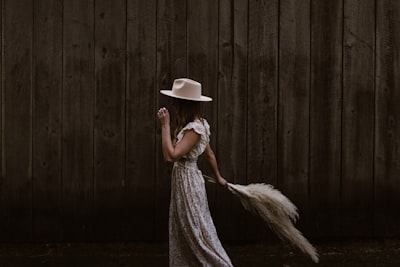 woman in white and brown floral dress wearing white hat standing beside brown wooden wall delicate teams background