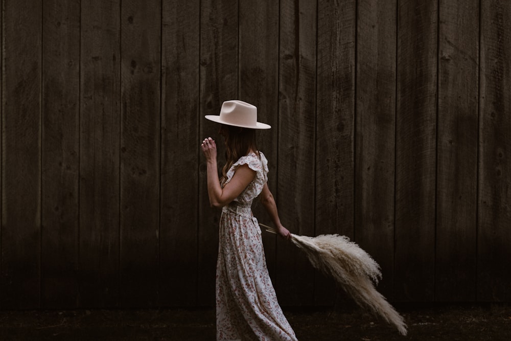 woman in white and brown floral dress wearing white hat standing beside brown wooden wall