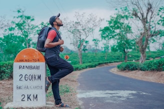 A person wearing a cap, sunglasses, and a backpack stands on one foot, leaning against a milestone on a rural road surrounded by green vegetation and trees. The milestone displays distances to nearby locations. The road curves into the distance with a backdrop of lush greenery under a clear sky.