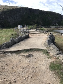 A rugged pathway made of concrete, framed by uneven stone formations, extends into a grassy landscape. Nearby, a small, white structure is visible against the backdrop of a steep rock face covered in green vegetation. A large wind turbine with blades is partially visible in the upper right corner under a patterned sky.