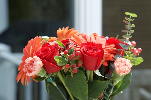 A vibrant bouquet featuring red roses, orange gerbera daisies, pink carnations, and green foliage. The arrangement is well-lit with a soft, natural background, highlighting the vivid colors of the flowers and leaves.