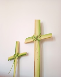 Two crosses made from palm leaves are displayed against a plain, off-white background. The larger cross has a knot tied at the intersection of the vertical and horizontal arms, with visible green and yellow shades. The smaller cross, also made from palm leaves, has a similar knot design and coloration.