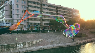 A large, iridescent soap bubble is being created with a triangular-shaped wand held by a person whose hand is visible. The bubble stretches across the image, with a backdrop of an urban area featuring a multi-story building and a walkway beside a body of water. The scene captures a bright, sunlit atmosphere with the sun setting in the background.