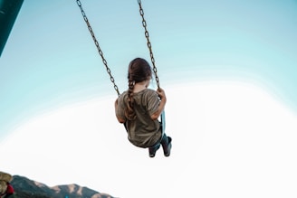 A child with a braid is swinging, facing away with the vast sky in the background, creating a sense of freedom and playfulness.