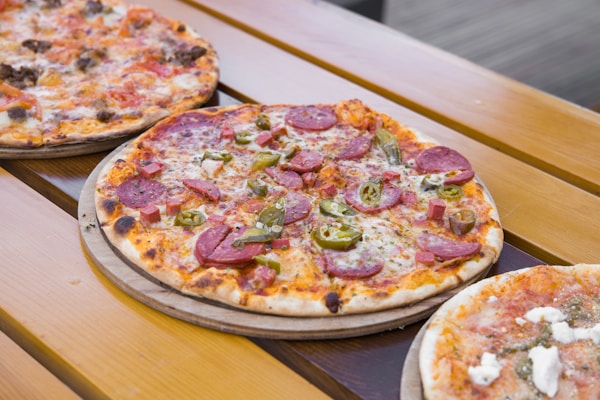 Three pizzas are placed on wooden boards outdoors. The pizza in the foreground features pepperoni slices, green jalapeños, and a layer of melted cheese. Adjacent pizzas appear to have varied toppings, with visible chunks of meat and white cheese. The setting suggests an informal dining atmosphere.