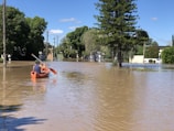A person in a canoe paddles through a flooded street surrounded by trees and utility poles. The water reflects the clear blue sky and nearby greenery, while buildings are partially submerged in the background.