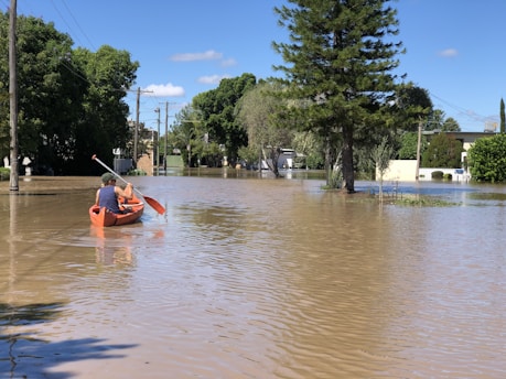 A person in a canoe paddles through a flooded street surrounded by trees and utility poles. The water reflects the clear blue sky and nearby greenery, while buildings are partially submerged in the background.