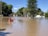 A person in a canoe paddles through a flooded street surrounded by trees and utility poles. The water reflects the clear blue sky and nearby greenery, while buildings are partially submerged in the background.