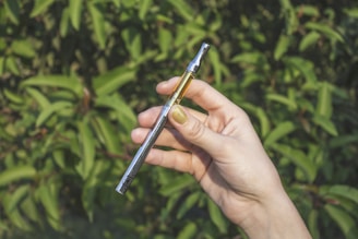 A hand with light skin is holding a vaping device, featuring a metal body and a transparent cartridge partially filled with liquid. The background consists of blurred green foliage.