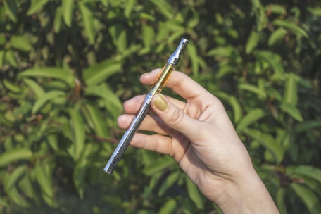 A hand with light skin is holding a vaping device, featuring a metal body and a transparent cartridge partially filled with liquid. The background consists of blurred green foliage.