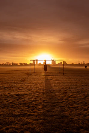 A silhouette of a young man kicking a soccer ball on an empty field during twilight.