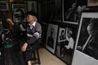 An elderly man wearing glasses and a beret is seated, holding a long paintbrush, surrounded by various framed black-and-white portraits. The walls are covered with these portraits, which include images of people in different poses and attire. The man appears to be painting or drawing, with tools and other art supplies scattered around.