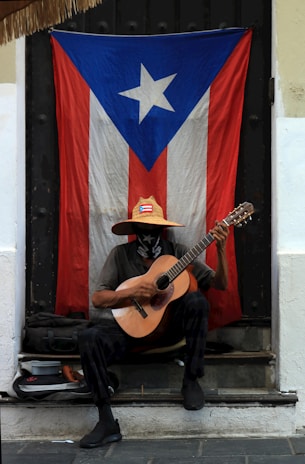 A person is sitting on a doorstep playing an acoustic guitar. They are wearing a hat with the Puerto Rican flag emblem on it and a mask. Behind them hangs a large Puerto Rican flag.