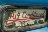 An old and weathered sign with the words 'Air Conditioned' in red cursive lettering. The sign's background is peeling, with visible wear and age, possibly on glass or an old vehicle window surrounded by a metallic frame. The visible blue paint suggests a retro or vintage aesthetic.