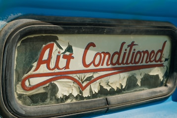 An old and weathered sign with the words 'Air Conditioned' in red cursive lettering. The sign's background is peeling, with visible wear and age, possibly on glass or an old vehicle window surrounded by a metallic frame. The visible blue paint suggests a retro or vintage aesthetic.