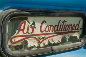 An old and weathered sign with the words 'Air Conditioned' in red cursive lettering. The sign's background is peeling, with visible wear and age, possibly on glass or an old vehicle window surrounded by a metallic frame. The visible blue paint suggests a retro or vintage aesthetic.