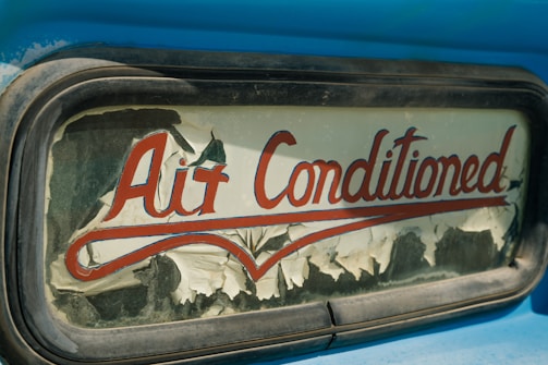 An old and weathered sign with the words 'Air Conditioned' in red cursive lettering. The sign's background is peeling, with visible wear and age, possibly on glass or an old vehicle window surrounded by a metallic frame. The visible blue paint suggests a retro or vintage aesthetic.