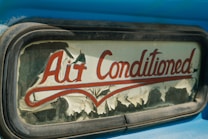 An old and weathered sign with the words 'Air Conditioned' in red cursive lettering. The sign's background is peeling, with visible wear and age, possibly on glass or an old vehicle window surrounded by a metallic frame. The visible blue paint suggests a retro or vintage aesthetic.