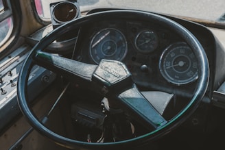 A close-up view of an old vehicle dashboard, featuring a large steering wheel with the Scania logo, several analog gauges including a speedometer, and a tachometer. The dashboard is worn, with visible scratches and dust, suggesting a vintage or well-used vehicle.