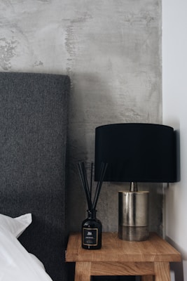 A modern bedroom setting featuring a dark grey upholstered headboard adjacent to a light wooden bedside table. On the table, there is a black diffuser bottle with reeds and a metallic lamp with a black shade. The backdrop consists of a textured grey concrete wall, creating an industrial aesthetic.