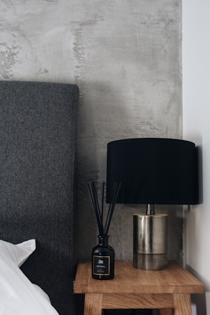A modern bedroom setting featuring a dark grey upholstered headboard adjacent to a light wooden bedside table. On the table, there is a black diffuser bottle with reeds and a metallic lamp with a black shade. The backdrop consists of a textured grey concrete wall, creating an industrial aesthetic.