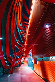 A futuristic building facade with sweeping red and orange curves illuminated by vibrant neon lighting. The architecture features bold lines and angles that create a dynamic and modern aesthetic. The pathway alongside the building is lit with multi-colored lights, enhancing the dramatic effect.