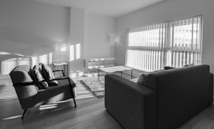 A modern living room with minimalistic furniture, including two armchairs, a sofa, a coffee table, and two standing lamps. The room is well-lit by natural light coming through large windows with blinds. The floor is wooden, and a small rug is placed between the seating area.