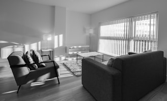 A modern living room with minimalistic furniture, including two armchairs, a sofa, a coffee table, and two standing lamps. The room is well-lit by natural light coming through large windows with blinds. The floor is wooden, and a small rug is placed between the seating area.
