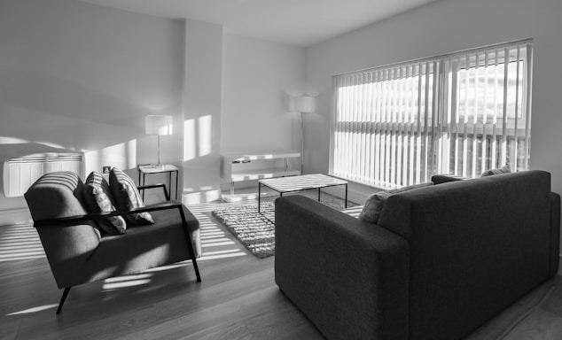 A modern living room with minimalistic furniture, including two armchairs, a sofa, a coffee table, and two standing lamps. The room is well-lit by natural light coming through large windows with blinds. The floor is wooden, and a small rug is placed between the seating area.
