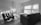 A modern living room with minimalistic furniture, including two armchairs, a sofa, a coffee table, and two standing lamps. The room is well-lit by natural light coming through large windows with blinds. The floor is wooden, and a small rug is placed between the seating area.