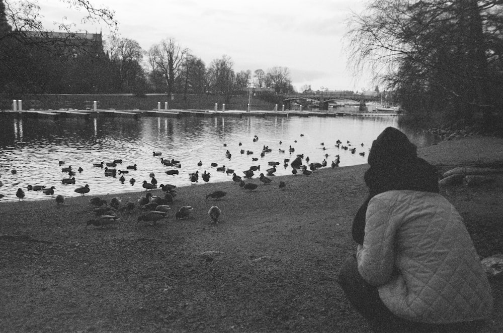 grayscale photo of woman sitting on grass field near lake