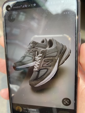 A person is holding a smartphone displaying a picture of gray athletic shoes with white laces. The phone screen shows details of a social media profile below the shoes.
