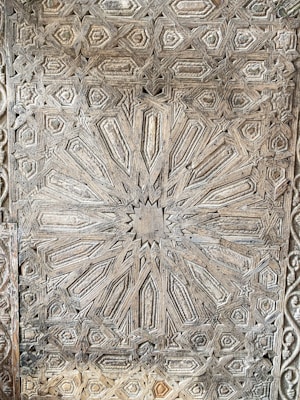 Intricately carved wooden panel featuring geometric patterns with a central starburst design. The craftsmanship highlights symmetrical shapes, possibly reflecting Islamic or Moorish architectural influences.