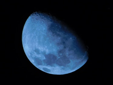 A close-up view of a half-moon with visible craters and surface textures, displayed in a cool blue hue against a stark black background.